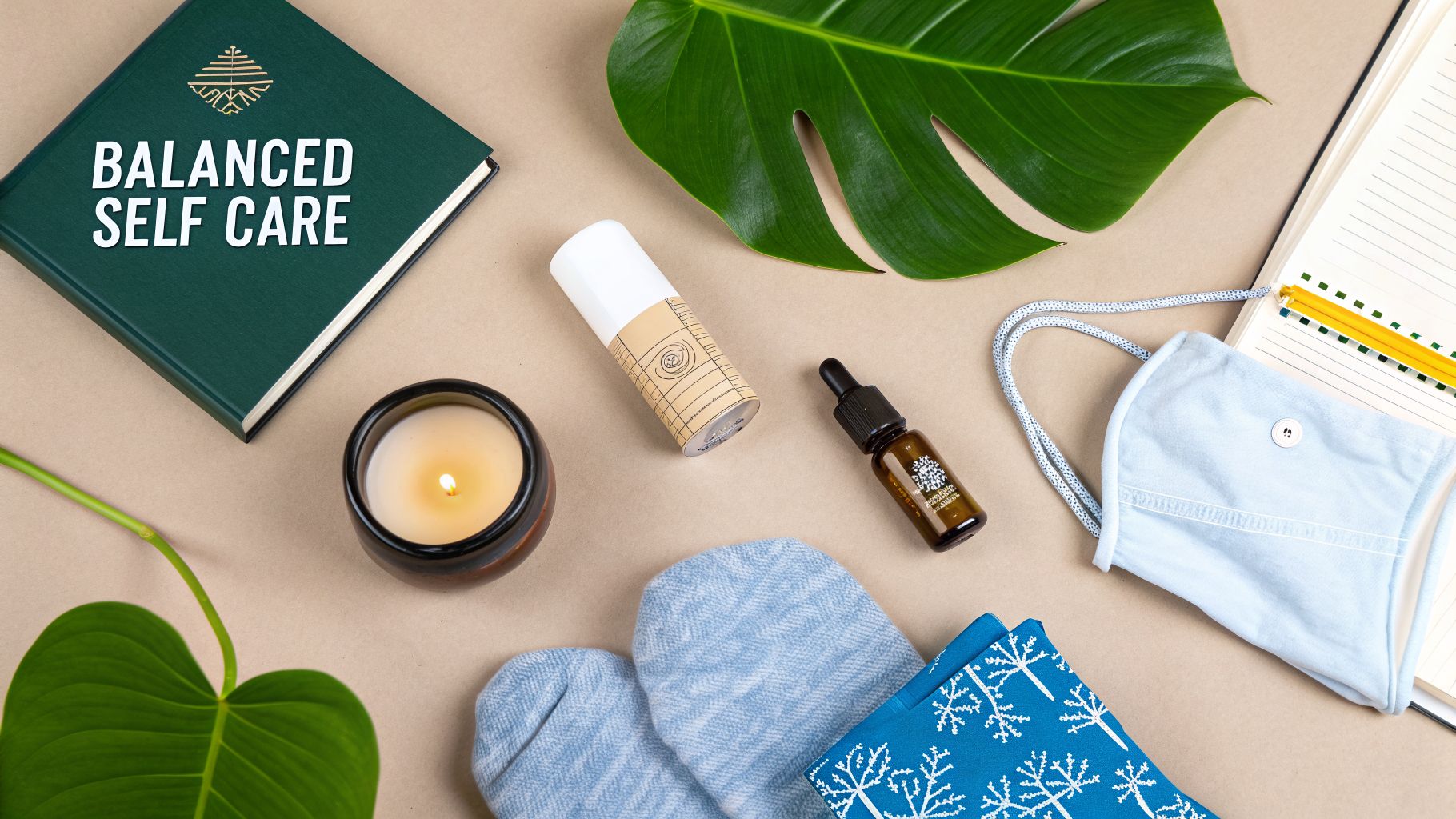 Overhead shot of self-care essentials: a 'Balanced Self Care' book, candle, oils, and cozy accessories on a beige surface.